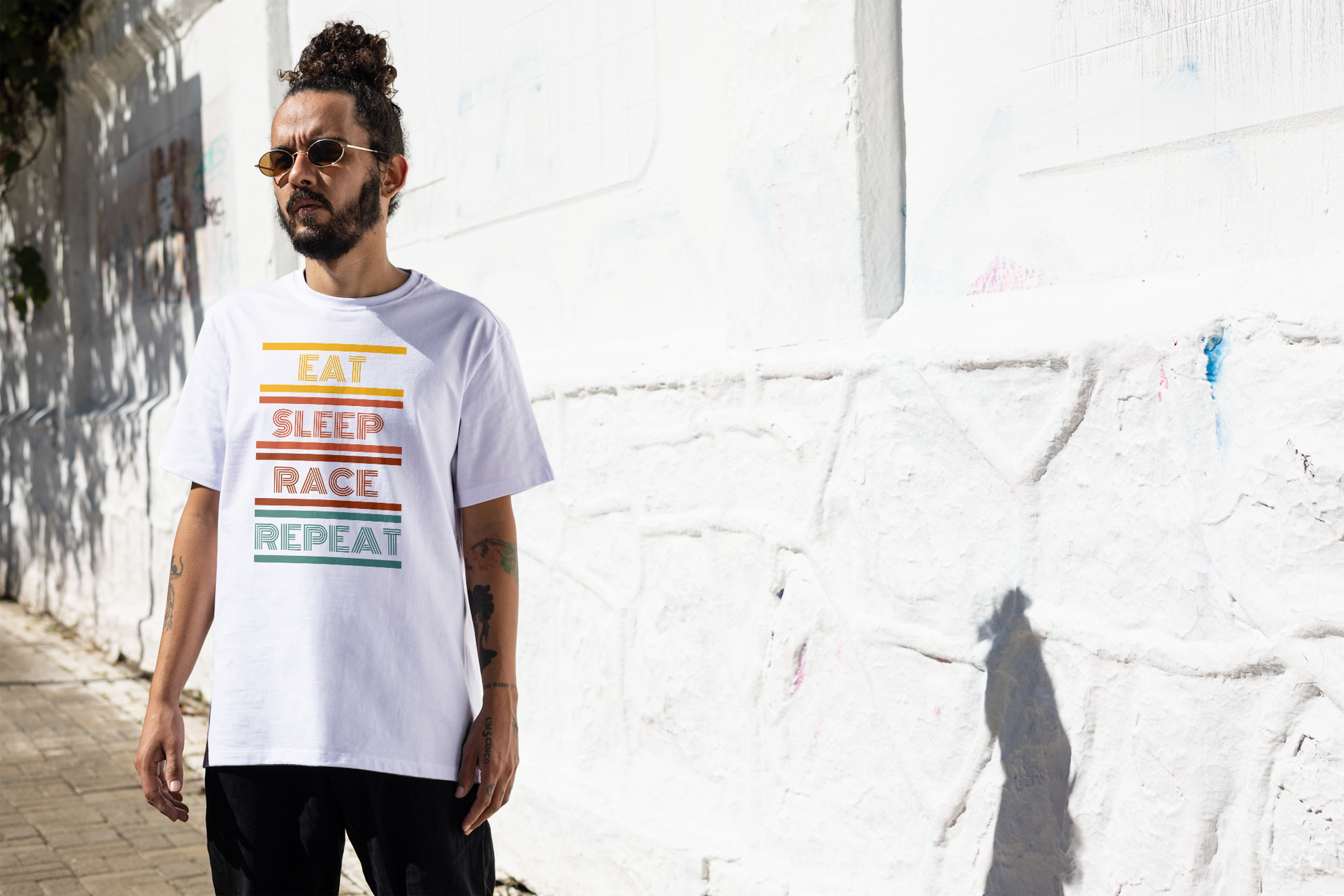 A man wearing a white graphic t-shirt with the words 'EAT SLEEP RACE REPEAT' in stacked horizontal stripes of yellow, orange, red and green, standing against a white textured wall.