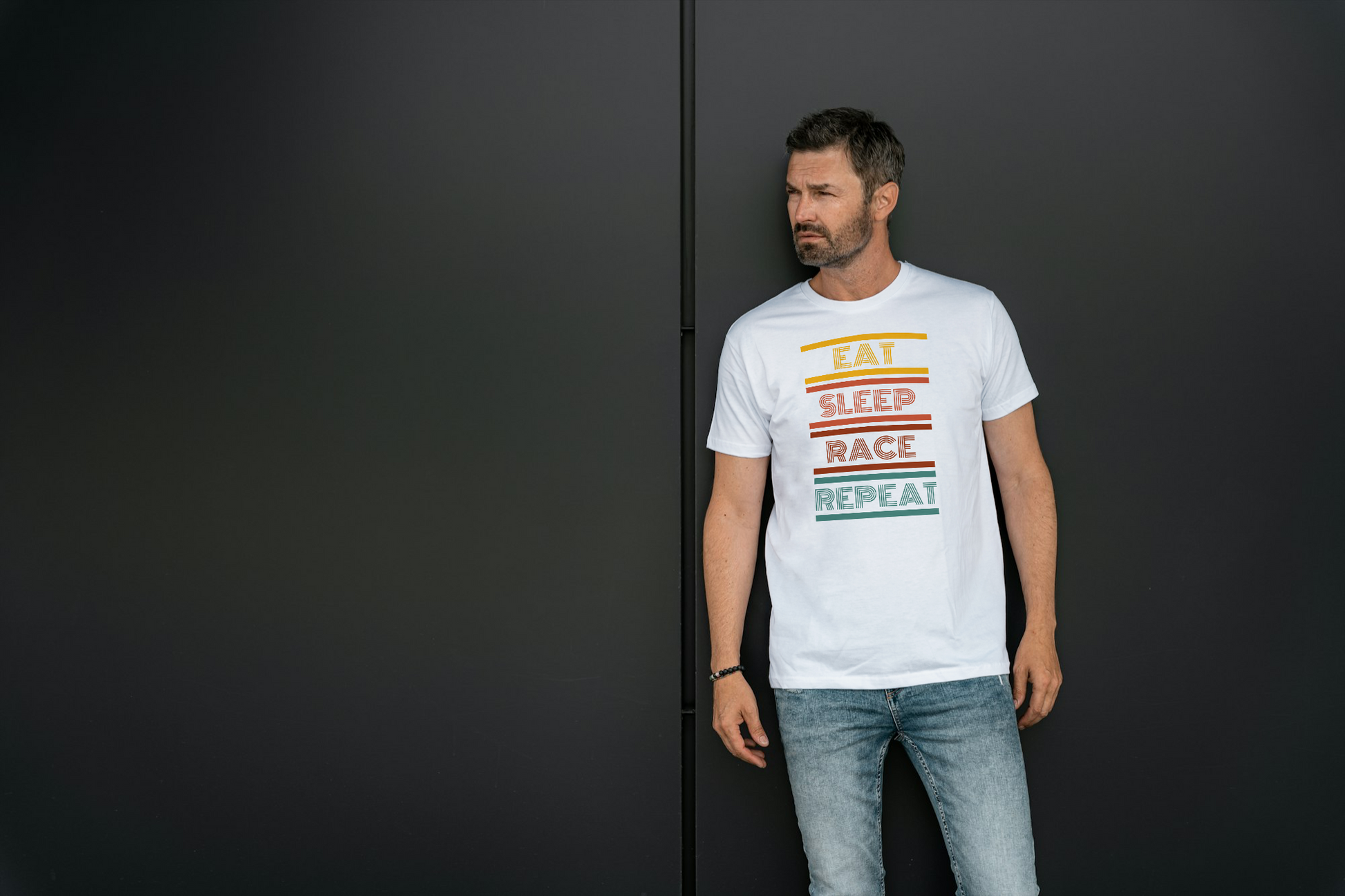 A man wearing a white graphic t-shirt with the words 'EAT SLEEP RACE REPEAT' in colorful stripes, standing against a dark gray wall.